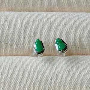 May include: A pair of small, green gourd-shaped stud earrings. The earrings have a silver-colored border and are set against a textured, off-white fabric background. The earrings are a bright, vibrant green.