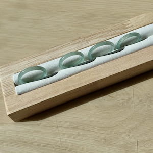 May include: Four light green jade rings displayed on a wooden jewelry holder with a white padded interior. The rings are translucent and have a smooth, polished finish. The wooden holder has a natural wood grain.