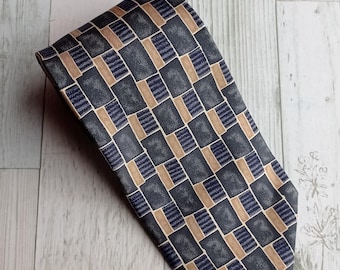 May include: A patterned necktie featuring a repeating geometric design. The tie's primary color is dark gray, with a pattern of squares and rectangles in tan and navy blue. The tie is folded and displayed on a light-colored surface.