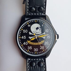 Men’s military quartz wristwatch inspired by the Spanish Army 8th Cavalry Regiment “Lusitania,” featuring a bold black dial with striking skull and crossbones emblem, a symbol of bravery, courage, and sacrifice. This collectible watch includes a precise quartz movement, durable black case, and a black leather strap with white stitching. Perfect for military history collectors, army enthusiasts, and men’s fashion lovers, this unworn timepiece represents unique heritage, bold design.