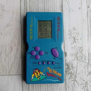 Close-up photo of a vintage WY-2007 Golden Light handheld console from the 1990s, featuring a blue body, purple buttons, and an LCD screen. The device offers 99 built-in games including Tetris. The packaging is aged, but the console itself is in new, unused condition. Perfect collectible for retro gaming fans and nostalgic 90s toy lovers.