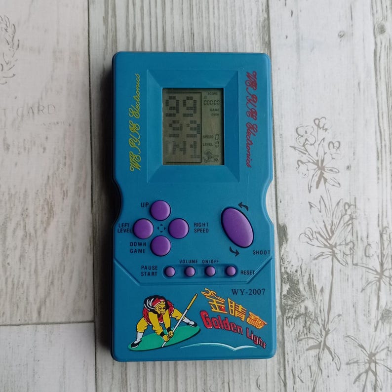Close-up photo of a vintage WY-2007 Golden Light handheld console from the 1990s, featuring a blue body, purple buttons, and an LCD screen. The device offers 99 built-in games including Tetris. The packaging is aged, but the console itself is in new, unused condition. Perfect collectible for retro gaming fans and nostalgic 90s toy lovers.