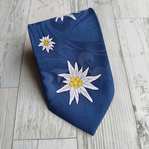 Può includere: Una cravatta blu scuro con un motivo ripetuto di fiori di stella alpina bianchi con centri gialli. Il tessuto presenta un sottile disegno tonale a vortice sullo sfondo, che conferisce un aspetto strutturato alla superficie blu profondo.