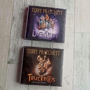 Puede incluir: Dos estuches de CD con novelas de Terry Pratchett. El estuche superior es para "Diggers" y el inferior para "Truckers". Ambos estuches tienen ilustraciones y el texto "Read by Tony Robinson".