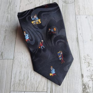 Puede incluir: Una corbata negra con un patrón repetido de figuras de dibujos animados. Las figuras visten de rojo, azul y amarillo, sobre un fondo gris oscuro con remolinos. La corbata está hecha de un material liso, posiblemente seda.