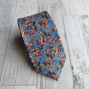 May include: A necktie featuring a light blue background with a dense, repeating pattern of small orange and brown flowers interspersed with green and brown leaves. The tie has a pointed tip and a smooth, flat surface texture throughout the design.