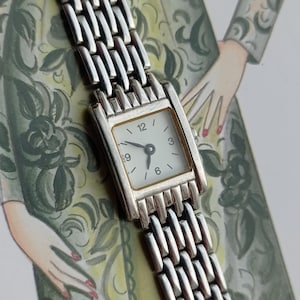 May include: A silver wristwatch with a rectangular face and a white dial. The watch has a linked metal strap. The watch face displays the numbers 12, 3, 6, and 9. The hands point to approximately 10:10.