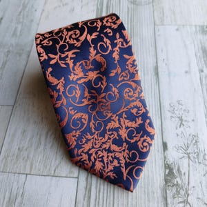 Pode incluir: Uma gravata azul marinho com um intrincado padrão floral laranja. A gravata é feita de um tecido liso e é exibida em uma superfície de madeira clara. O design apresenta folhas e vinhas em redemoinho.