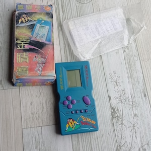 Close-up photo of a vintage WY-2007 Golden Light handheld console from the 1990s, featuring a blue body, purple buttons, and an LCD screen. The device offers 99 built-in games including Tetris. The packaging is aged, but the console itself is in new, unused condition. Perfect collectible for retro gaming fans and nostalgic 90s toy lovers.