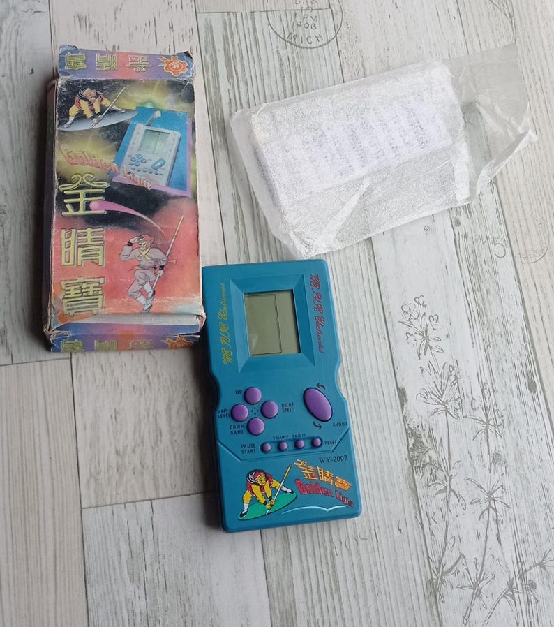 Close-up photo of a vintage WY-2007 Golden Light handheld console from the 1990s, featuring a blue body, purple buttons, and an LCD screen. The device offers 99 built-in games including Tetris. The packaging is aged, but the console itself is in new, unused condition. Perfect collectible for retro gaming fans and nostalgic 90s toy lovers.