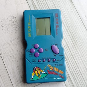 May include: A teal handheld electronic game console with a small screen and purple buttons. The game has the text "WE PYIE Electronics" and "Golden Light" with a graphic of a person on a surfboard. The model number is WY-2007.