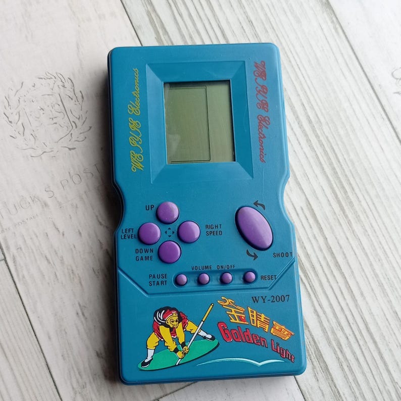 Close-up photo of a vintage WY-2007 Golden Light handheld console from the 1990s, featuring a blue body, purple buttons, and an LCD screen. The device offers 99 built-in games including Tetris. The packaging is aged, but the console itself is in new, unused condition. Perfect collectible for retro gaming fans and nostalgic 90s toy lovers.