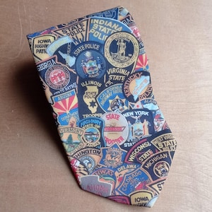 May include: A patterned necktie featuring various state police and highway patrol badges. The tie is predominantly navy blue with colourful patches from different states, including Georgia, New York, and California.