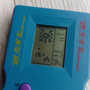 Close-up photo of a vintage WY-2007 Golden Light handheld console from the 1990s, featuring a blue body, purple buttons, and an LCD screen. The device offers 99 built-in games including Tetris. The packaging is aged, but the console itself is in new, unused condition. Perfect collectible for retro gaming fans and nostalgic 90s toy lovers.
