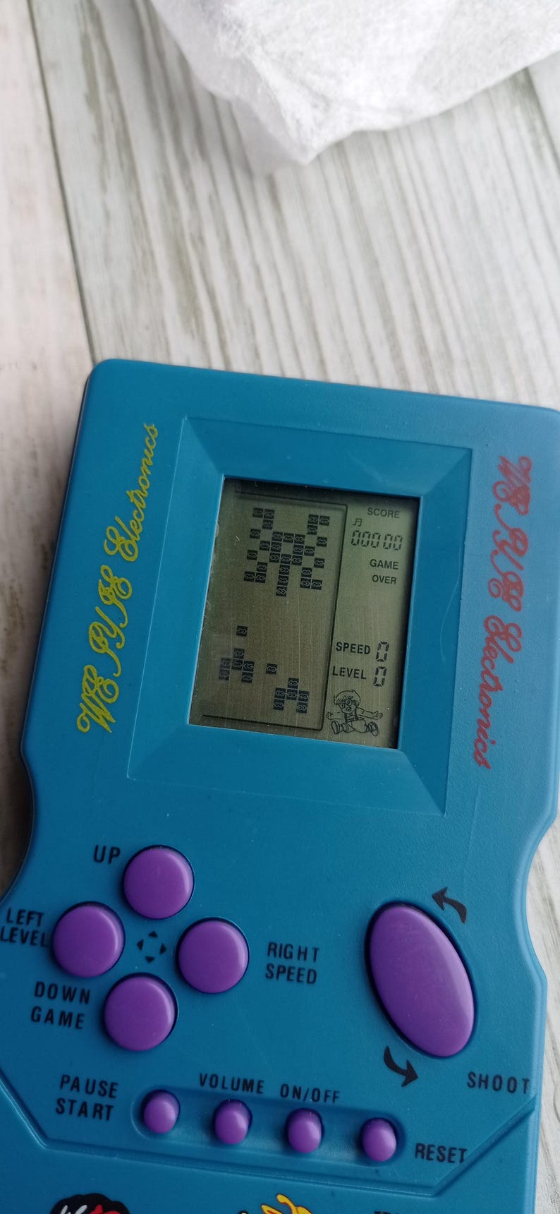 Close-up photo of a vintage WY-2007 Golden Light handheld console from the 1990s, featuring a blue body, purple buttons, and an LCD screen. The device offers 99 built-in games including Tetris. The packaging is aged, but the console itself is in new, unused condition. Perfect collectible for retro gaming fans and nostalgic 90s toy lovers.
