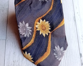 May include: A dark blue necktie with a floral pattern. The tie features yellow and white flowers with orange wavy lines. The tie is made of a textured fabric and is displayed on a light wood surface.