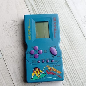 Close-up photo of a vintage WY-2007 Golden Light handheld console from the 1990s, featuring a blue body, purple buttons, and an LCD screen. The device offers 99 built-in games including Tetris. The packaging is aged, but the console itself is in new, unused condition. Perfect collectible for retro gaming fans and nostalgic 90s toy lovers.