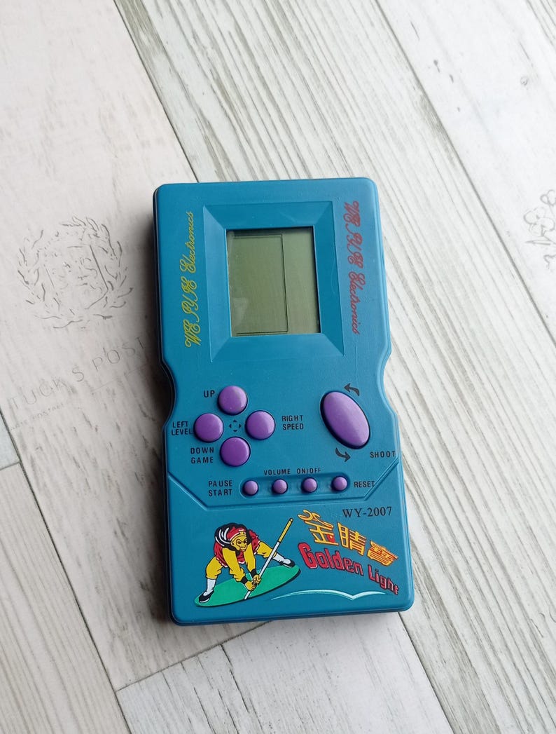 Close-up photo of a vintage WY-2007 Golden Light handheld console from the 1990s, featuring a blue body, purple buttons, and an LCD screen. The device offers 99 built-in games including Tetris. The packaging is aged, but the console itself is in new, unused condition. Perfect collectible for retro gaming fans and nostalgic 90s toy lovers.