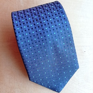 Op de afbeelding: Een marineblauwe stropdas met een geweven patroon van donkerblauwe vierkanten en turquoise diamantaccenten. Het ontwerp van de stropdas vervaagt van een dicht patroon aan de bovenkant naar een spaarzamer patroon aan de onderkant. De stropdas is van zijde.
