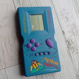 Close-up photo of a vintage WY-2007 Golden Light handheld console from the 1990s, featuring a blue body, purple buttons, and an LCD screen. The device offers 99 built-in games including Tetris. The packaging is aged, but the console itself is in new, unused condition. Perfect collectible for retro gaming fans and nostalgic 90s toy lovers.