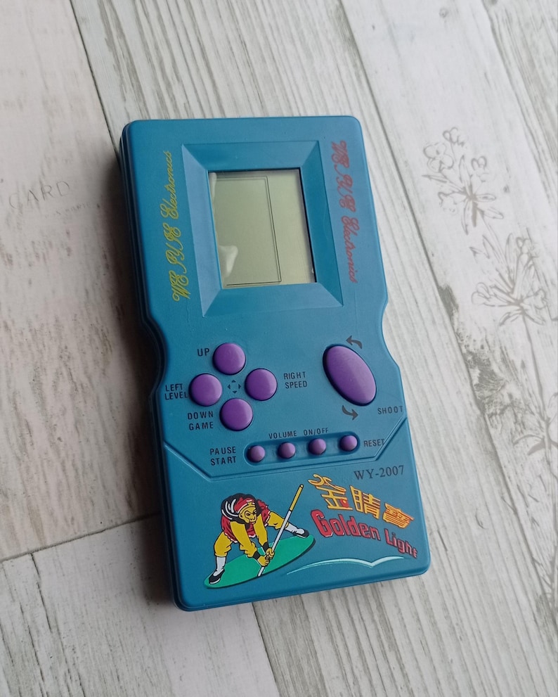 Close-up photo of a vintage WY-2007 Golden Light handheld console from the 1990s, featuring a blue body, purple buttons, and an LCD screen. The device offers 99 built-in games including Tetris. The packaging is aged, but the console itself is in new, unused condition. Perfect collectible for retro gaming fans and nostalgic 90s toy lovers.