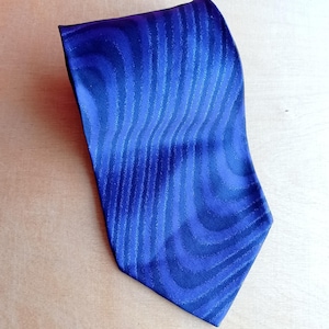 Può includere: Una cravatta blu con un motivo a righe ondulate. La cravatta è di una tonalità blu intenso con strisce blu più chiare che creano un aspetto strutturato. La cravatta è realizzata in un materiale lucido.