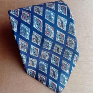 May include: A navy blue tie featuring a repeating diamond pattern. Each diamond contains a paisley design in blue, red, and beige. The tie is made of a smooth fabric, suitable for formal or business wear. The tie is approximately 147 cm long.