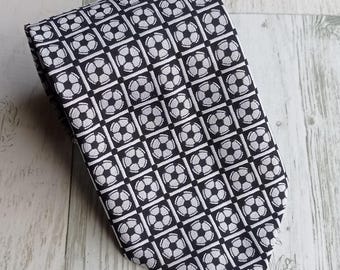 May include: A black and white patterned necktie featuring a repeating soccer ball design. The tie is made of a woven fabric and is suitable for formal or casual wear. The soccer ball pattern is in a grid layout.