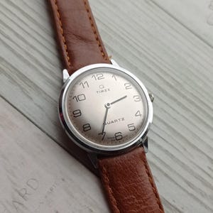 Könnte beinhalten: Eine silberfarbene Timex Q Quartz-Uhr mit einem braunen Lederarmband. Das Zifferblatt hat schwarze Ziffern und Zeiger, mit dem Markennamen und "QUARTZ". Die Uhr liegt vor einem hellen Holzmaserungshintergrund.