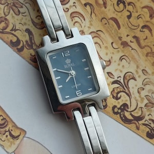 May include: A silver-toned Royal London wristwatch with a square, blue face. The watch features the numbers 12 and 6, and the words "ROYAL" and "LONDON" are printed on the face. The watch band is also silver-toned.