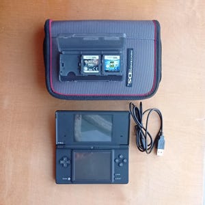 May include: A black Nintendo DS handheld console with a grey carry case. The case holds two game cartridges. A USB charging cable is also included. The console is on a wooden surface.