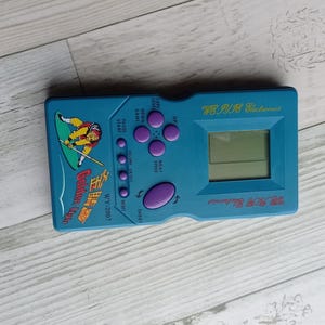 Close-up photo of a vintage WY-2007 Golden Light handheld console from the 1990s, featuring a blue body, purple buttons, and an LCD screen. The device offers 99 built-in games including Tetris. The packaging is aged, but the console itself is in new, unused condition. Perfect collectible for retro gaming fans and nostalgic 90s toy lovers.