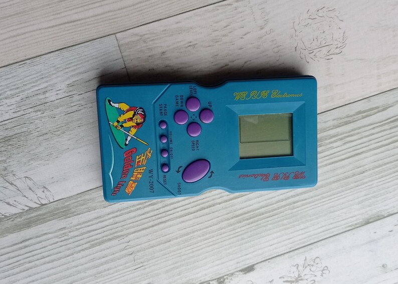 Close-up photo of a vintage WY-2007 Golden Light handheld console from the 1990s, featuring a blue body, purple buttons, and an LCD screen. The device offers 99 built-in games including Tetris. The packaging is aged, but the console itself is in new, unused condition. Perfect collectible for retro gaming fans and nostalgic 90s toy lovers.