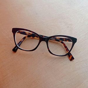 May include: Tortoiseshell-patterned eyeglasses with a cat-eye design. The glasses have a black frame and clear lenses. The brand name "MARC JACOBS" is visible on the frame. The glasses are on a light brown wooden surface.