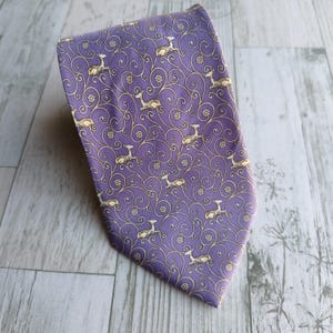 Könnte beinhalten: Eine lila Krawatte mit einem sich wiederholenden Muster aus Hirschen und floralen Designs in Gold. Die Krawatte ist aus einem glatten Stoff gefertigt und ist zur unteren rechten Ecke hin abgewinkelt. Der Hintergrund ist eine hellfarbige Holzoberfläche.