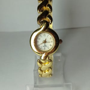 May include: A gold-tone wristwatch with a white face and black Roman numeral hour markers. The watch has a gold-tone metal band with a unique link design. The watch face has the text "JC PARIS" printed on it.