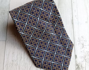 Könnte beinhalten: Eine marineblaue Krawatte mit einem sich wiederholenden geometrischen Muster aus goldenen, grauen und weißen Quadraten. Die Krawatte ist gefaltet und liegt auf einer hellen Holzoberfläche. Das Muster erzeugt ein gitterartiges Design.