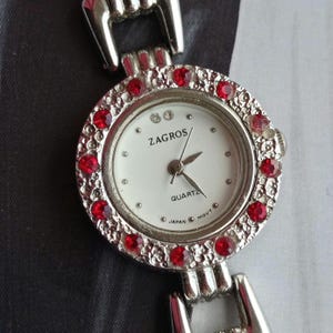May include: A silver Zagros quartz watch with a white face and red gemstone detailing. The watch has a round face with a silver bezel and a silver strap. The watch face displays the word "Zagros".