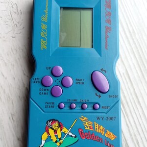 Close-up photo of a vintage WY-2007 Golden Light handheld console from the 1990s, featuring a blue body, purple buttons, and an LCD screen. The device offers 99 built-in games including Tetris. The packaging is aged, but the console itself is in new, unused condition. Perfect collectible for retro gaming fans and nostalgic 90s toy lovers.
