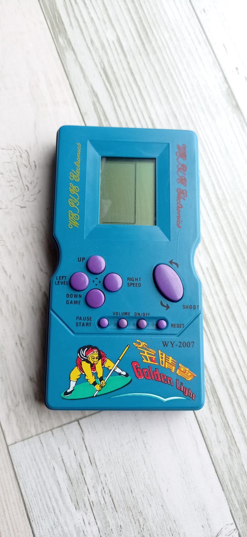 Close-up photo of a vintage WY-2007 Golden Light handheld console from the 1990s, featuring a blue body, purple buttons, and an LCD screen. The device offers 99 built-in games including Tetris. The packaging is aged, but the console itself is in new, unused condition. Perfect collectible for retro gaming fans and nostalgic 90s toy lovers.