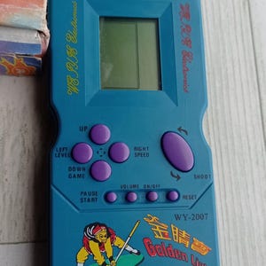 Close-up photo of a vintage WY-2007 Golden Light handheld console from the 1990s, featuring a blue body, purple buttons, and an LCD screen. The device offers 99 built-in games including Tetris. The packaging is aged, but the console itself is in new, unused condition. Perfect collectible for retro gaming fans and nostalgic 90s toy lovers.