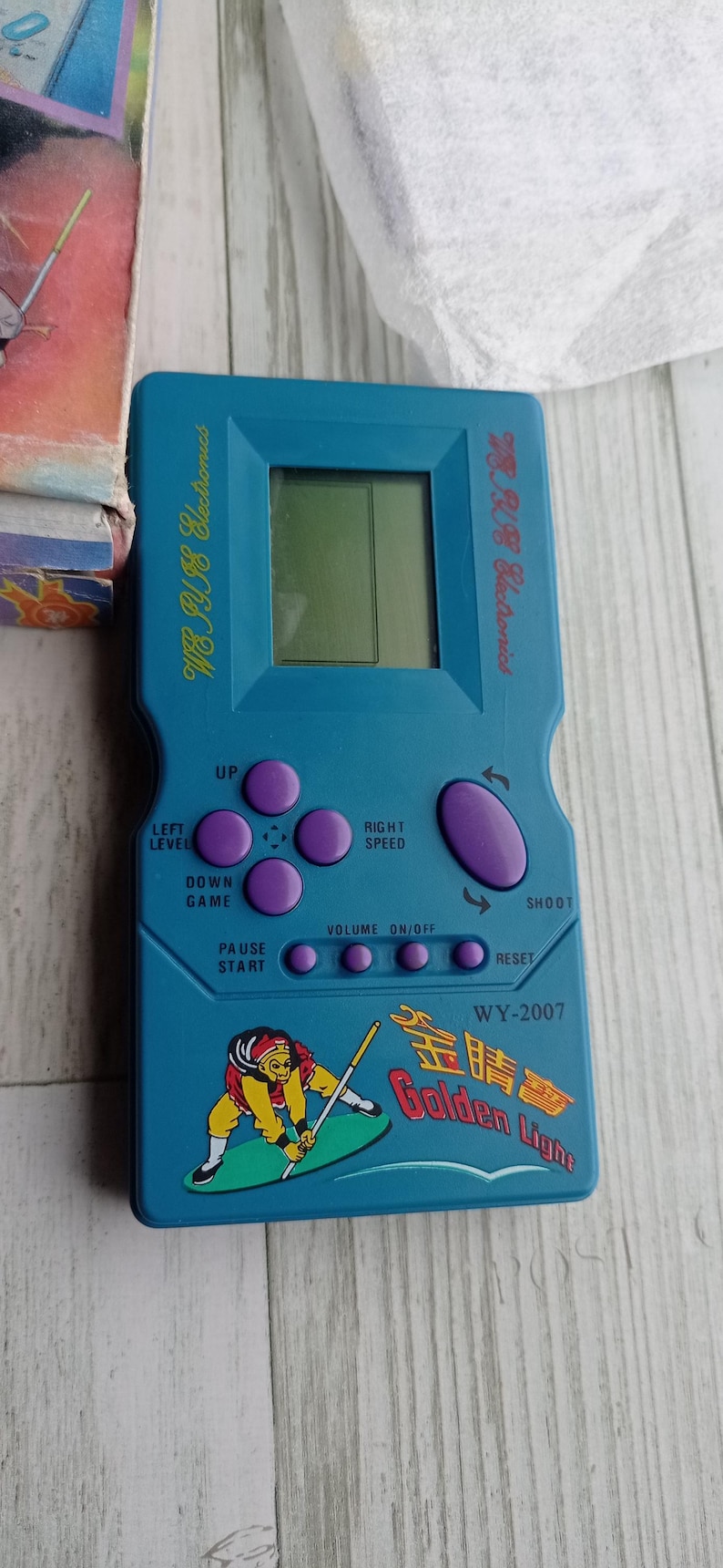 Close-up photo of a vintage WY-2007 Golden Light handheld console from the 1990s, featuring a blue body, purple buttons, and an LCD screen. The device offers 99 built-in games including Tetris. The packaging is aged, but the console itself is in new, unused condition. Perfect collectible for retro gaming fans and nostalgic 90s toy lovers.