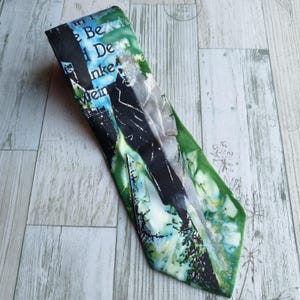May include: A necktie with a unique abstract design. The tie features a blend of green, black, and blue hues with areas of text. The tie is made of a smooth fabric and is displayed on a light wood surface.