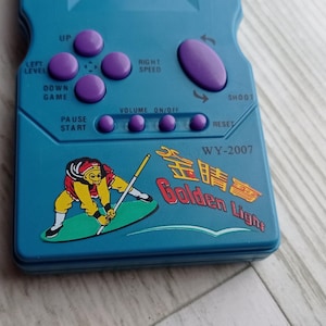 Close-up photo of a vintage WY-2007 Golden Light handheld console from the 1990s, featuring a blue body, purple buttons, and an LCD screen. The device offers 99 built-in games including Tetris. The packaging is aged, but the console itself is in new, unused condition. Perfect collectible for retro gaming fans and nostalgic 90s toy lovers.