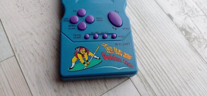 Close-up photo of a vintage WY-2007 Golden Light handheld console from the 1990s, featuring a blue body, purple buttons, and an LCD screen. The device offers 99 built-in games including Tetris. The packaging is aged, but the console itself is in new, unused condition. Perfect collectible for retro gaming fans and nostalgic 90s toy lovers.