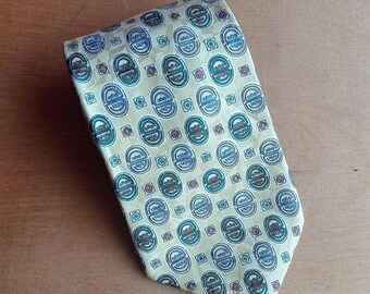 May include: A yellow tie with a repeating pattern of green, blue, and red circular designs. The tie is folded and lies on a wooden surface. The pattern is evenly spaced across the fabric.
