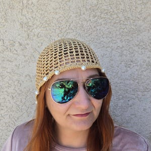 May include: A beige crocheted hat with a grid pattern and white pearl accents. The hat is worn with aviator sunglasses with a reflective lens. The person is wearing a light purple t-shirt.