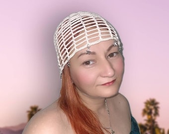 May include: A white crocheted head covering with a grid-like pattern. The head covering is adorned with small, silver-colored star charms. The background features a sunset with palm trees.