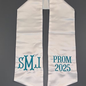 May include: White satin graduation sash with teal embroidery. The left side features the initials "SMJ", and the right side reads "PROM 2025". The sash is designed to be worn over the shoulders.