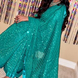 May include: A teal-colored sari with sequin embellishments. The fabric is draped, creating flowing lines and a shimmering effect. The sari is a vibrant teal color, with a hint of blue. The garment is worn with a matching teal blouse.